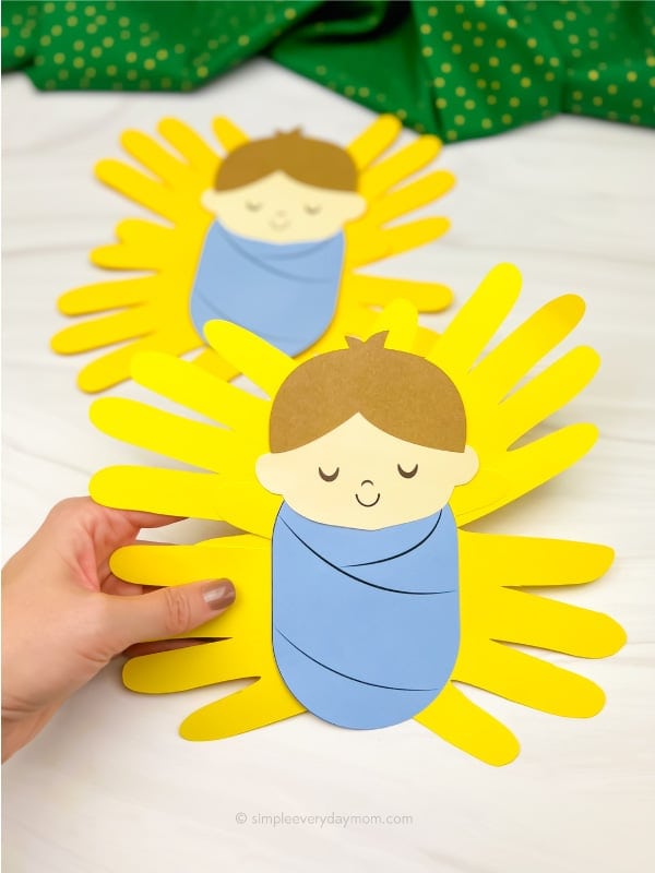 Colorful paper craft of a baby wrapped in a blue blanket with yellow handprint rays, perfect for nursery decor or baby shower gift.