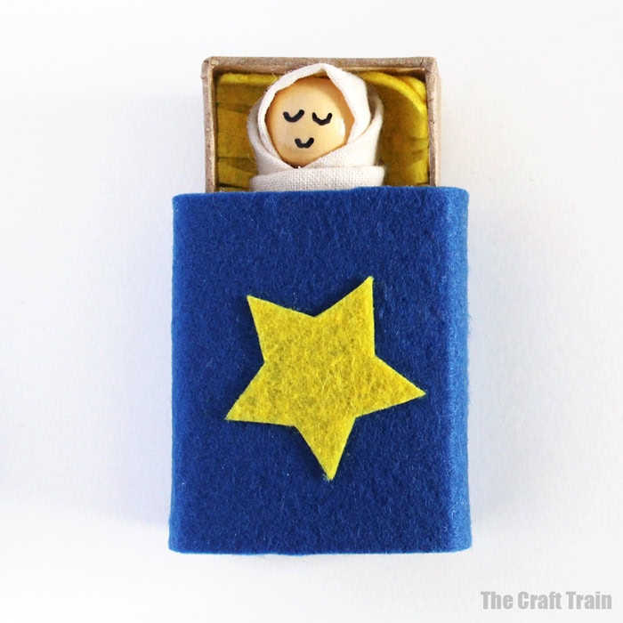 Adorable handmade baby doll with a smiling face, wrapped in a white blanket, resting in a blue felt bed with a yellow star, perfect for nursery decor or gift.