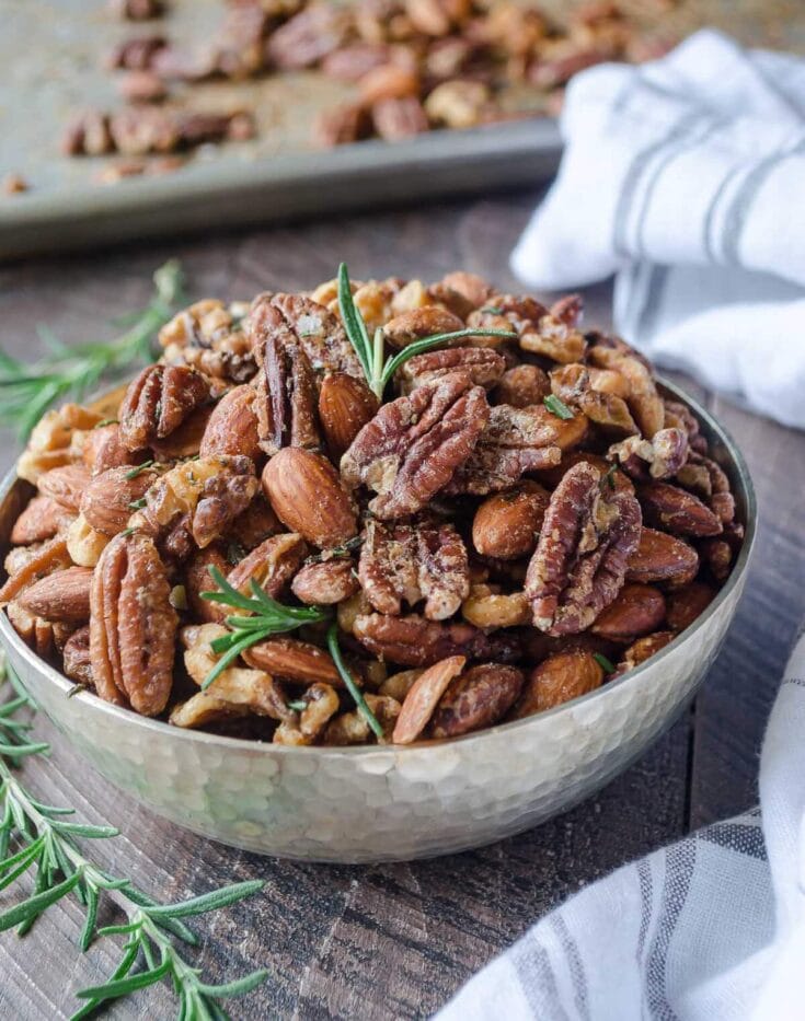 A bowl of roasted mixed nuts including pecans, almonds, and walnuts garnished with fresh rosemary on a rustic wooden surface.