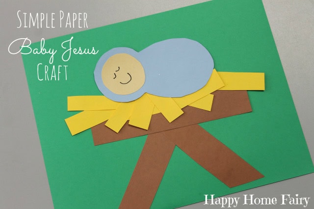Colorful paper craft of Baby Jesus lying on a yellow manger with a blue blanket, on a green background, for Christmas and religious holiday decorations.