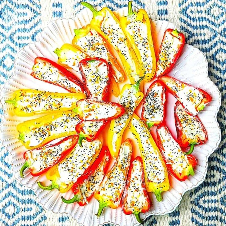 A vibrant platter of stuffed bell peppers filled with cheese and herbs, garnished with poppy seeds, on a decorative plate with a blue patterned cloth background.