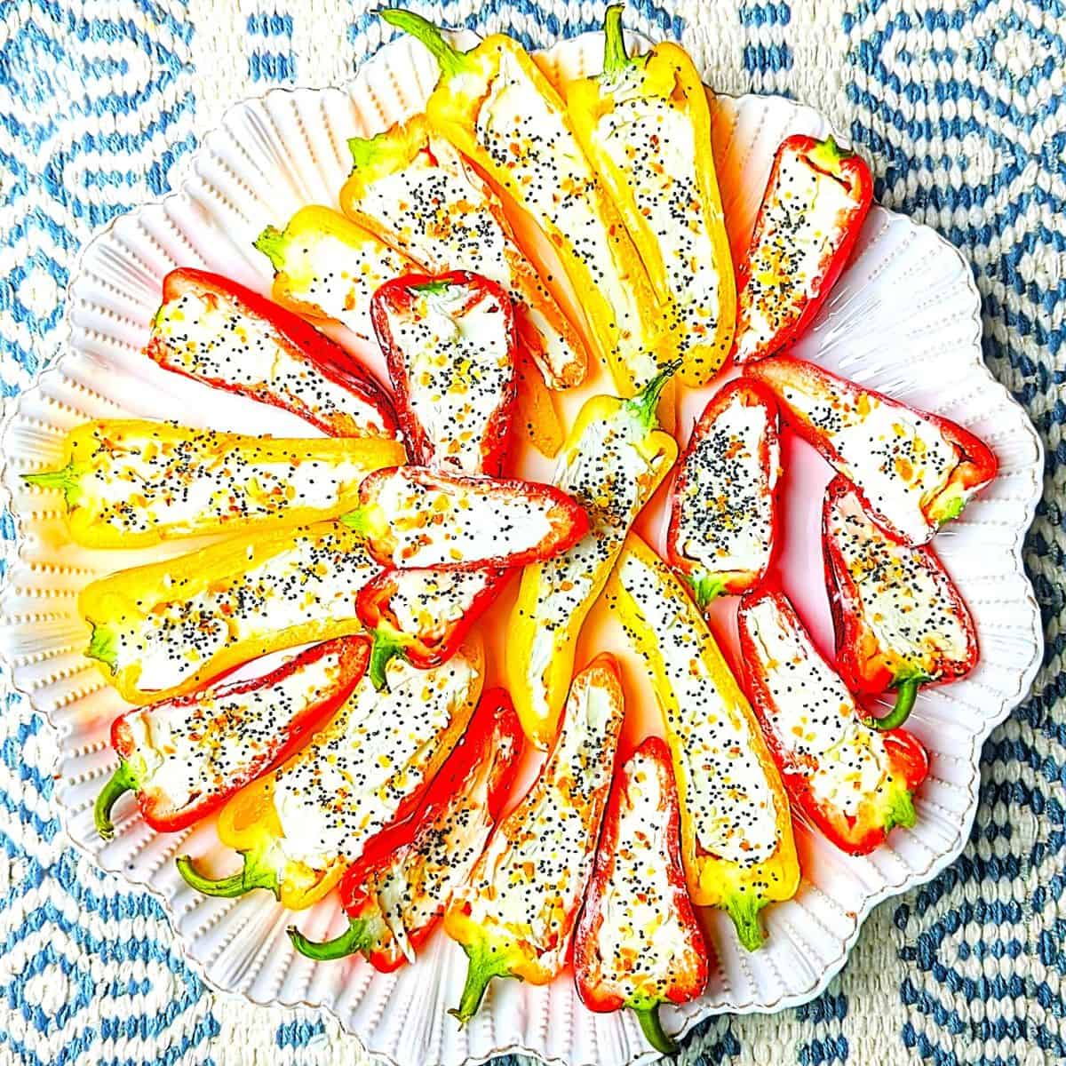 A vibrant platter of stuffed bell peppers filled with cheese and herbs, garnished with poppy seeds, on a decorative plate with a blue patterned cloth background.