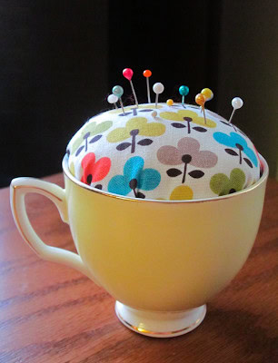 A decorative pin cushion with colorful pins inside a yellow teacup, perfect for sewing or craft projects, on a wooden surface.