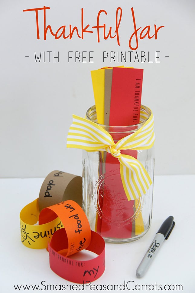 A glass jar decorated with a yellow ribbon contains colorful paper notes for gratitude, surrounded by additional paper strips and a black marker, on a white background.