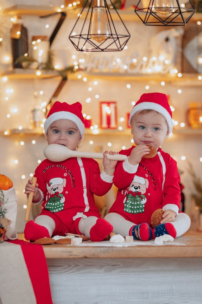 Two young children dressed in festive Christmas pajamas and Santa hats, sitting on a wooden surface with holiday decorations and fairy lights in the background.