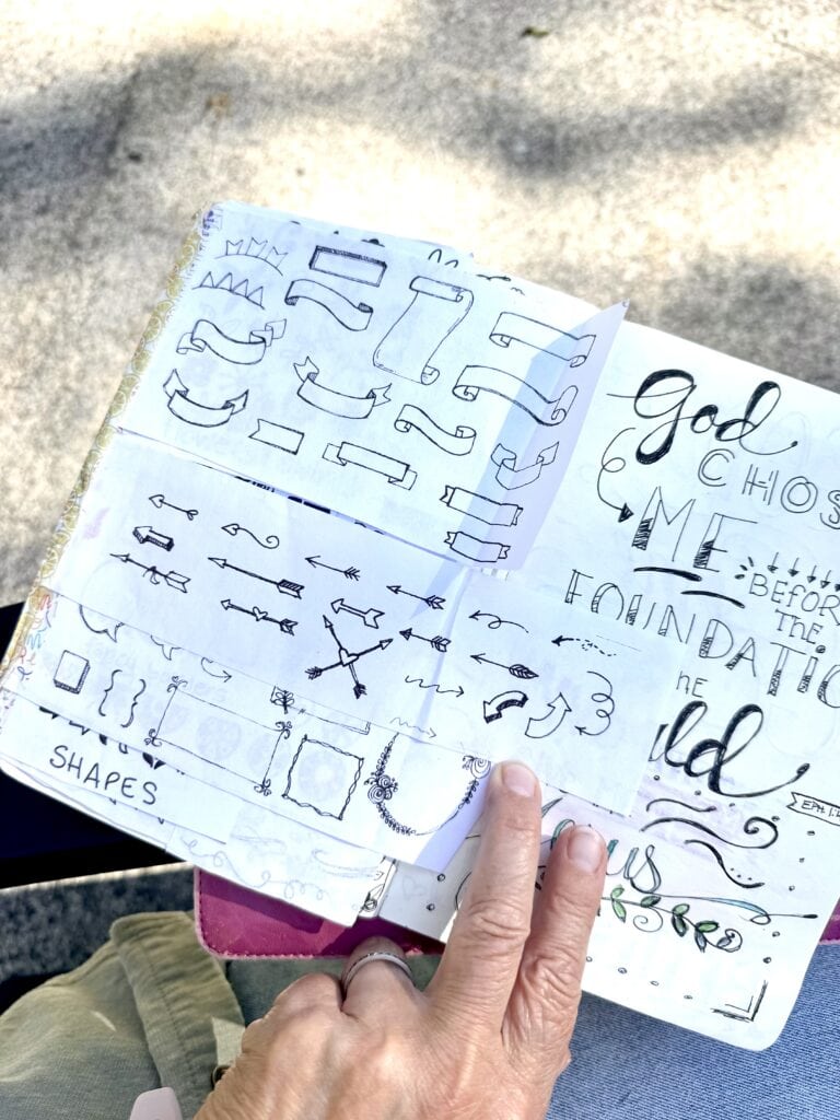 Open sketchbook with hand-drawn lettering and design sketches, held by a person outdoors, showcasing artistic typography and creative lettering ideas.