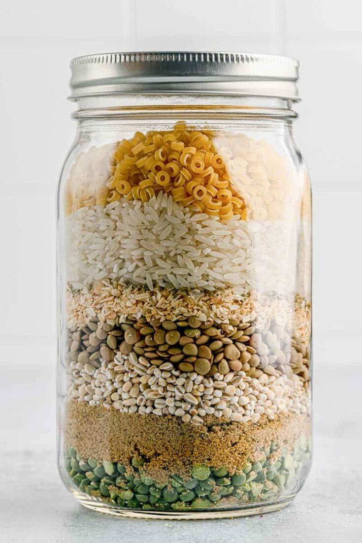 Assorted dry pantry ingredients layered in a glass mason jar, including pasta, rice, seeds, grains and spices for a colorful, organized kitchen display.