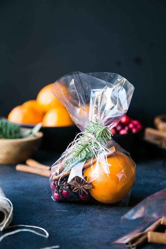 Ornamental gift bag with bright oranges, wrapped in clear cellophane and tied with rosemary sprig and cinnamon sticks on a dark surface, styled for handmade gift presentation.
