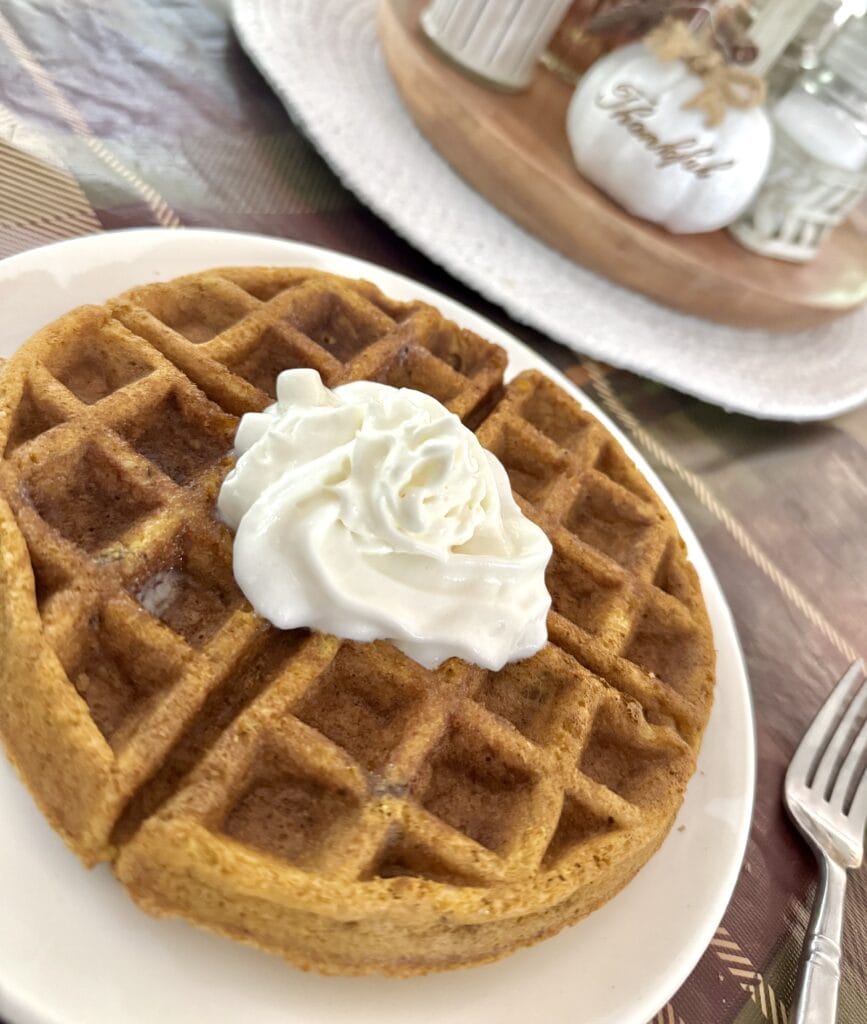 Close-up of a golden-brown waffle topped with a dollop of whipped cream on a white plate, with a cozy breakfast setting in the background.