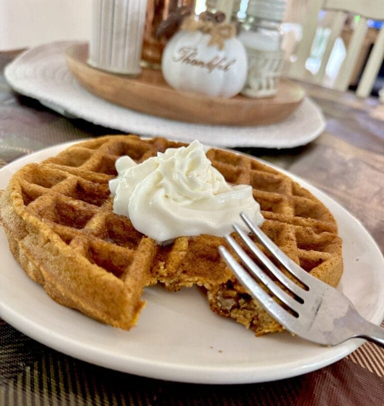 Close-up of a golden-brown waffle topped with whipped cream on a white plate, with a cozy kitchen background featuring decorative pumpkins and a 'Thankful' sign.