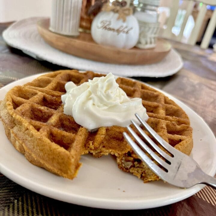 Close-up of a golden-brown waffle topped with whipped cream on a white plate, with a cozy kitchen background featuring decorative pumpkins and a 'Thankful' sign.