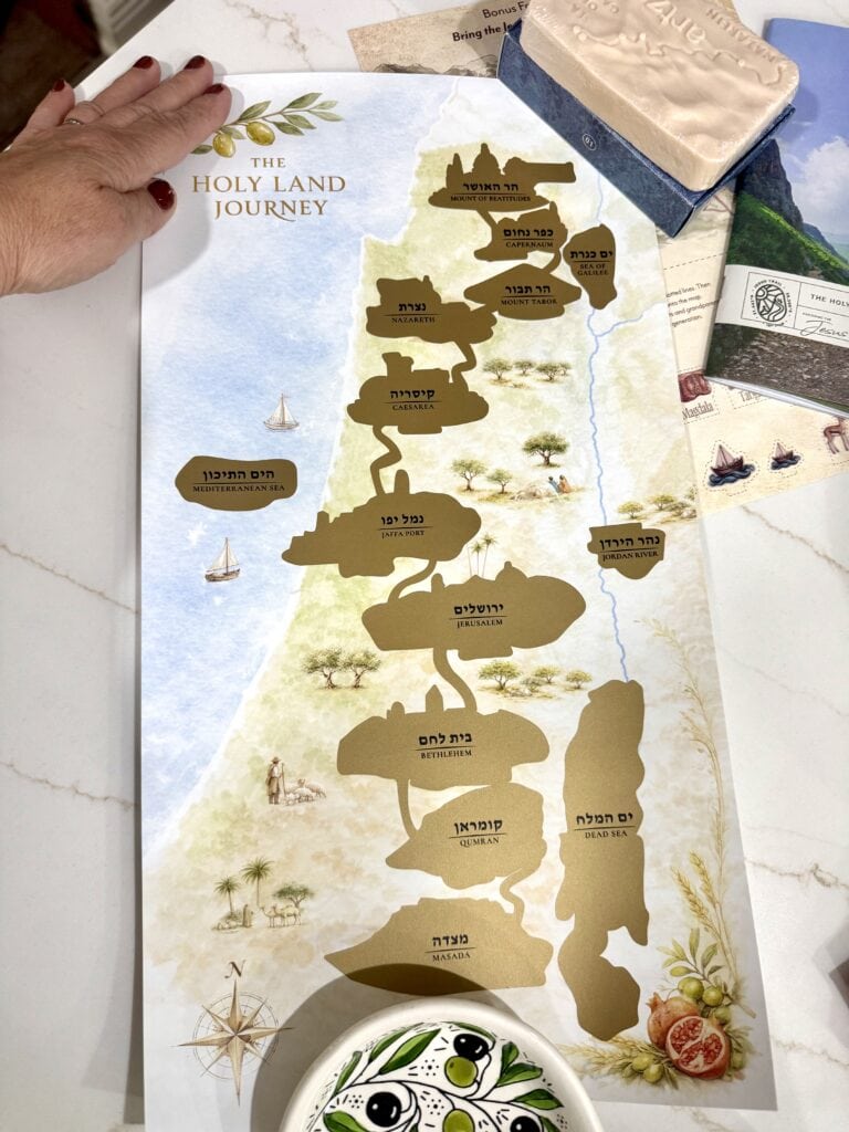 A map of the Holy Land Journey with labeled regions and a hand pointing to the map, accompanied by a pouch and booklets on a light marble surface 
