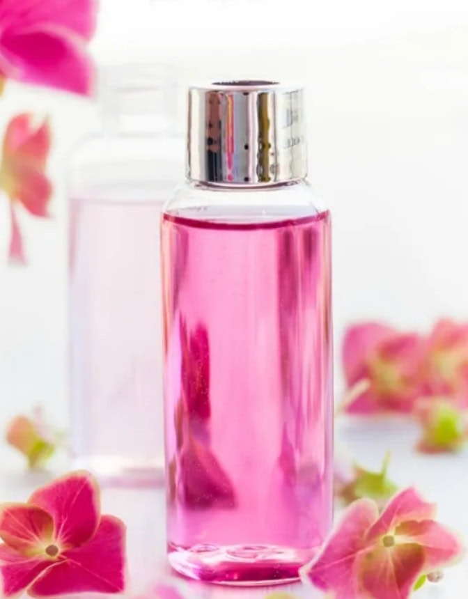 Pink perfume bottle with silver cap surrounded by soft pink flowers, bright white background, aesthetic fragrance photo for gift ideas and homemade care products.