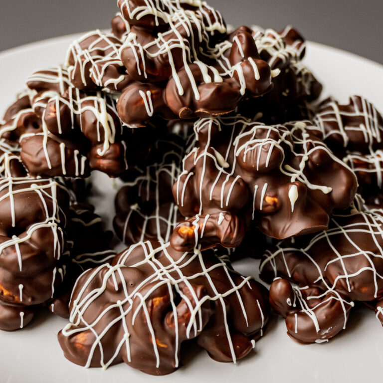Close-up of glossy chocolate clusters with white drizzle, peanut or nut centers, arranged on a white plate for holiday indulgence.