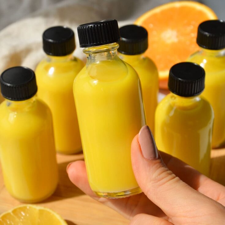 Close-up of a hand holding a small glass bottle filled with yellow lemon juice, surrounded by several similar bottles with black caps and a sliced orange in the background, on a wooden surface.