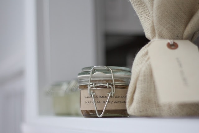 A small glass jar labeled natural body scrub sits on a white shelf beside a beige burlap-wrapped package, creating a calm, spa-like aesthetic suitable for a Hundred Affections product photo.