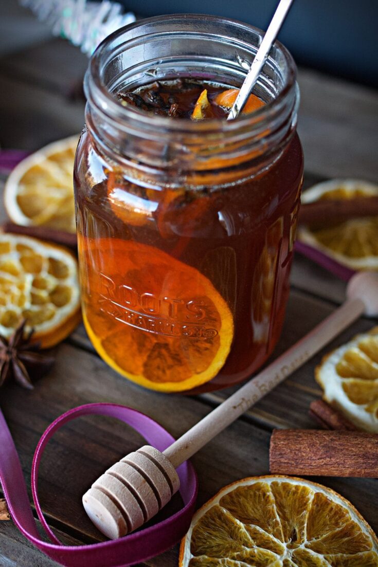 Jar of iced tea with orange slices, lemon slices, cinnamon sticks, and a honey dipper on a rustic wooden table.